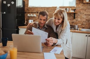 A couple are in a kitchen reading their post, the man is sat down and the woman is behind him and they are both reading the same letter 