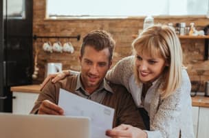 A man is sitting at a table, and a woman is leaning over his shoulder as they read a letter in the kitchen.